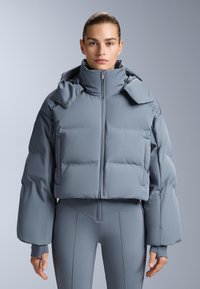 Skraćena puffer jakna u svijetlosivoj boji, sa visokim ovratnikom, podstavljenim dijelovima i bočnim džepovima. Glatka tekstura s detaljima koje se pričvršćuju na ramenima.