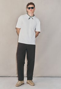 Vit kortärmad t-shirt med ett grönt mönstrat bandana, svarta löst sittande byxor och beige skor med texturerad sula, mot en neutral bakgrund.