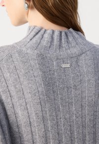 Šedý žebrovaný svetr s vysokým límcem, s vertikálními pruhy a malou kovovou nášivkou na zadní straně. Měkká textura a neformální design.