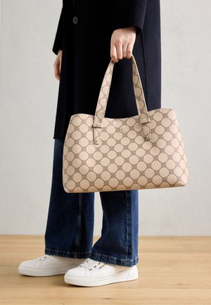 Personne portant un manteau bleu marine et un jean tenant un sac à main beige à motifs, debout sur un sol en bois, portant des baskets blanches.