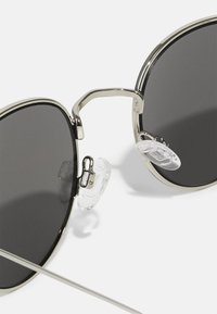 Occhiali da sole rotondi con lenti grigio scuro, montatura in metallo argentato e naselli regolabili trasparenti. Presentano un design elegante e minimalista.