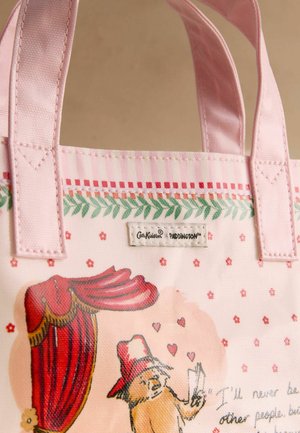 Vaaleanpunainen kangaskassi kangaskahvoilla, jossa on Paddington-nalle -kuvitus, jossa nalle pitää kirjettä punaisen verhon alla, jonka yksityiskohtina on kukkia ja sydämiä.