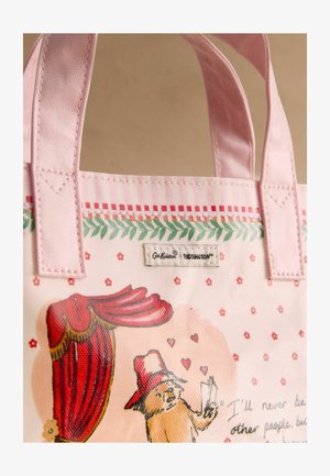 Roosa kangast käekott, millel on kangast sangad ja Paddington Beari illustratsioon, kus ta hoiab punase kardina all kirjaga ning millel on lille- ja südamekujundused.