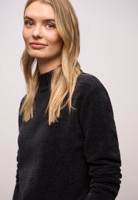 Czarny teksturowany sweter z wysokim kołnierzem i długimi rękawami. Gładka tkanina z subtelnym wzorem, noszony przez modelkę z długimi włosami.