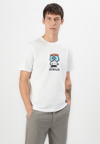 Joven con el cabello corto que lleva una camiseta blanca con un personaje de dibujos animados y la palabra "GENIO," combinada con pantalones grises.