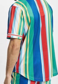 Monivärinen lyhythihainen paita, jossa pystysuuntaiset raidat punaisena, vihreänä, sinisenä, keltaisena ja valkoisena sekä hihansuissa resorikoristeet.