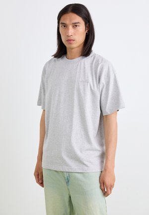 T-shirt en coton gris à manches courtes, col rond et petit logo sur la poitrine. Porté avec un pantalon vert clair. Texture lisse.