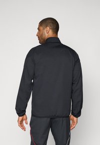 Veste légère noire avec un col haut, texture lisse et poignets élastiques. La vue arrière montre une coupe décontractée et un design minimal.