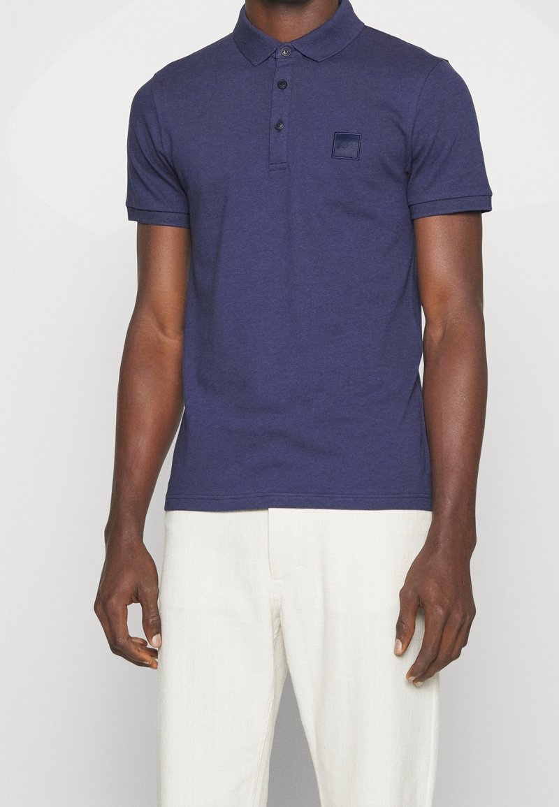 Homme portant un polo bleu marine à manches courtes avec col et un pantalon blanc, debout devant un fond clair uni.