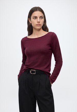 Femme aux cheveux châtain foncé portant un haut bordeaux à manches longues, un pantalon noir taille haute et une ceinture noire, debout les mains dans les poches.