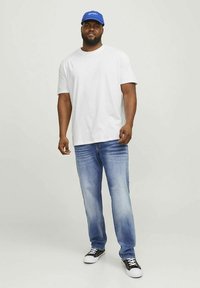Wit T-shirt, blauwe pet met tekst, lichtblauwe spijkerbroek, zwarte sneakers met witte zolen. Casual outfit, staande tegen een neutrale achtergrond.