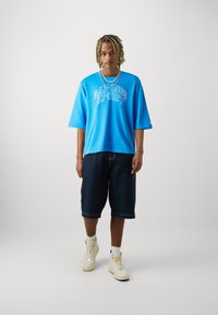 T-shirt azul oversize com a impressão "GAP 1969 SPORT", combinado com calções de ganga escuros até ao joelho e ténis de cano alto bejes.