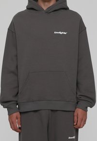 Grijze hoodie gemaakt van zachte stof, met een voorzak, geribde manchetten en een klein wit logo op de borst. Past bij grijze joggingbroek.