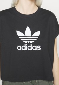 Black cropped t-shirt with white Adidas trefoil logo and text, worn by a person with light skin and blonde hair.