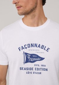 Camiseta de algodón blanca con texto gráfico en azul marino, que presenta un diseño de bandera y coordenadas costeras, cuello redondo y mangas cortas.