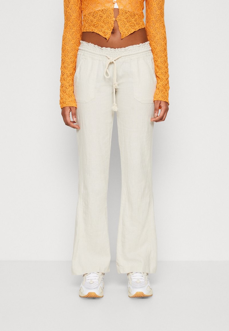 Kobieta ubrana w kremowe spodnie z troczkami, białe sneakersy i pomarańczowy teksturowany krótki kardigan na jednolitym tle.