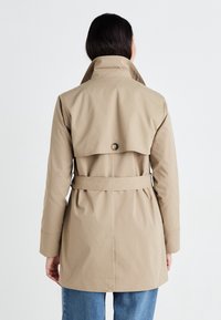 Beżowy płaszcz trench z wysokim kołnierzem, z paskiem w talii i tylnym klapem. Miękka faktura z zapięciem na guziki z tyłu. Zaprojektowany w luźnym fasonie.