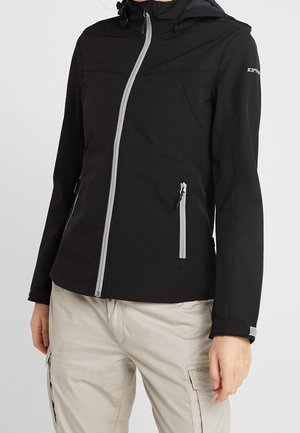 Person, die eine schwarze Kapuzenjacke mit grauem Reißverschluss und beige Cargohosen trägt, steht vor einem schlichten Hintergrund.