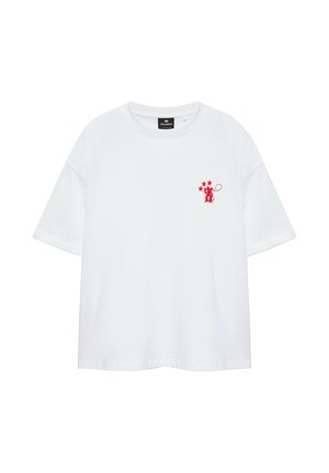 Mladý muž stojí s rukami v kapse, má na sebe biele voľné tričko s červeným logom, voľné svetlomodré rifle a hnedé žabky na jednoduchom pozadí.