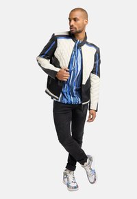 Vatteret jakke i sort og creme med blå accenter, parret med en mønstret blå skjorte, sorte jeans og multicolor sneakers.