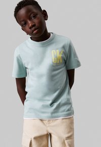 Camiseta de manga corta en verde menta con un destacado logo amarillo "CK" en el pecho, combinada con pantalones cortos cargo beige. Textura suave.