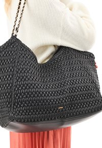 Personne portant un pull blanc et une jupe corail, portant un grand sac à bandoulière en cuir tressé noir avec une chaîne.