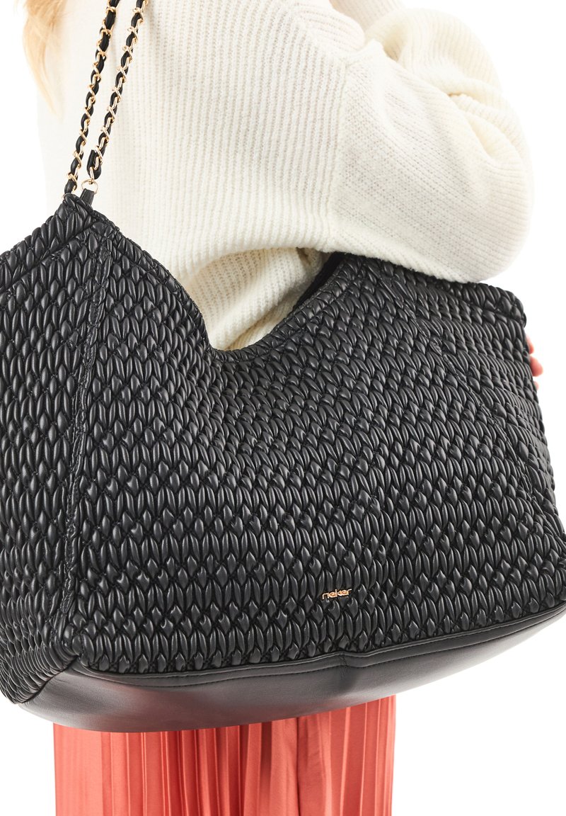 Personne portant un pull blanc et une jupe corail, portant un grand sac à bandoulière en cuir tressé noir avec une chaîne.