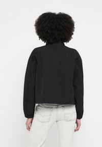 Melnā cropped jaciņa ar augstu apkakli, apjomīgām piedurknēm un gludu tekstūru, kombinēta ar gaišām džinsa biksēm.