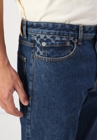 Jeans i denim i mörkblått med en mönstrad fickdetalj. Har kontrasterande sömmar och knappstängning. Materialet verkar mjukt och något texturerat.