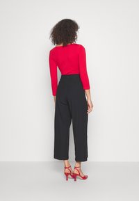 Červený top s dlouhým rukávem, který je zkombinován s černými širokými kalhotami s vysokým pasem a červenými lodičkami na vysokém podpatku, zobrazený ze zadní strany na bílém pozadí.