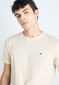 Camiseta de algodón beige con escote redondo y mangas cortas, que presenta un pequeño logo bordado en el lado izquierdo del pecho. Textura suave.