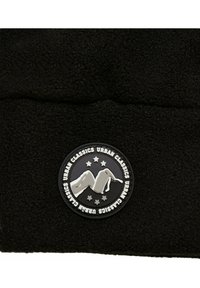Must mustri kangas, millel on ümmargune must ja hõbedane logo, kus on mägikujutis ja tähed, ning mille ääres on silt "URBAN CLASSICS".