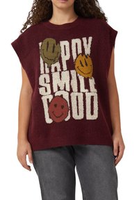 Ärmelloser kastanienbrauner Pullover mit großen weißen Buchstaben "HAPPY SMILE LOUD" und drei bunten lächelnden Gesichtern, getragen mit grauen Hosen.