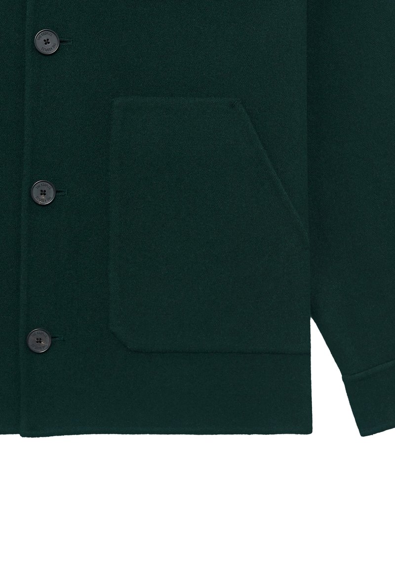 Mørkegrøn uldfrakke med sorte knapper og en stor skrå lomme fortil nederst til venstre, der viser en del af et manchet-sleeve.