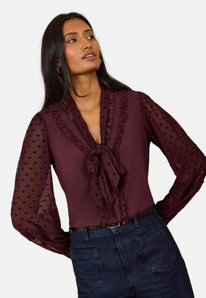 Kvinde iført en gennemsigtig, langærmet bordeauxbluse med sløjfe ved halsen og flæsedetaljer, kombineret med mørkeblå jeans med høj talje.