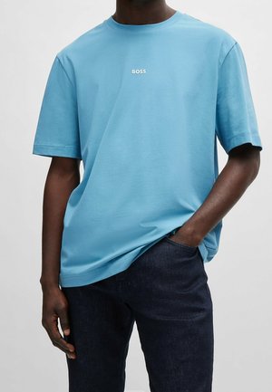Homme portant un t-shirt bleu clair avec le texte "BOSS" sur la poitrine et un jean bleu foncé, une main dans la poche, sur fond uni.