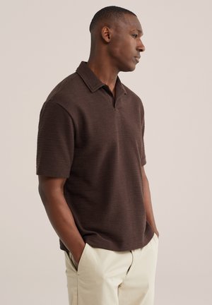 Čovjek stoji bočno s rukama u džepovima, nosi tamnosmeđu teksturiranu polo majicu kratkih rukava i svijetlo bež hlače.