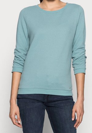 Mujer con un top de manga larga texturizado azul claro con escote redondo y jeans azul oscuro, de pie con los brazos relajados a los lados.