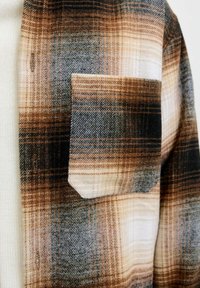 Brown and beige plaid shirt with a textured finish, featuring a chest pocket and a button closure. The pattern includes varying shades and lines.