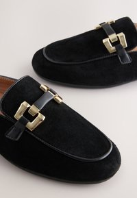Mocassins en daim noir avec une bordure en cuir lisse et des accents de quincaillerie dorée. Bout arrondi et silhouette élégante.