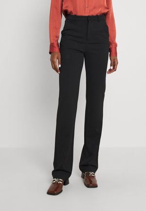 Persoon die hoge zwarte broek, roestkleurige blouse met lange mouwen en bruine hakken met metalen gespen draagt, staand op een grijze vloer.