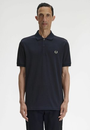 Man met kort donker haar die een zwart polohemd met korte mouwen draagt, met een klein wit laurierkranslogo op de borst.