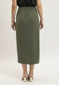 Jupe mi-longue, verte olive, avec une ceinture élastique, en tissu doux et texturé, et à coupe droite. Associée à des sandales à talons carrés argentées.