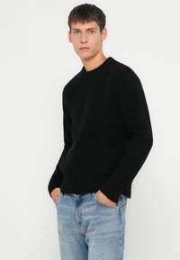 Zwarte geribbelde breisweater met een ronde hals, lange mouwen en een ontspannen pasvorm, gecombineerd met lichtblauwe jeans.