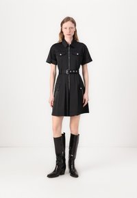 Vestido preto de mangas curtas com gola, fecho éclair frontal, dois bolsos no peito e cintura com cinto. Combinado com botas altas pretas brilhantes até aos joelhos.