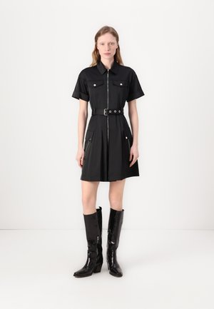 Robe noire à manches courtes avec un col, fermeture éclair à l'avant, deux poches poitrine et une taille ceinturée. Assortie à des bottes noires brillantes montantes.