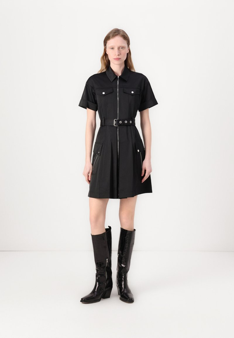 Vestido preto de mangas curtas com gola, fecho éclair frontal, dois bolsos no peito e cintura com cinto. Combinado com botas altas pretas brilhantes até aos joelhos.