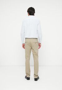 Man wearing beige trousers, a light blue checkered long-sleeve shirt, and black shoes, standing facing a white wall.