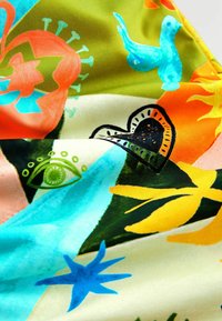 Kleurige stof met abstracte ontwerpen: een blauwe vogel, een groene oog, een hartvorm en verschillende bladeren in oranje, geel en turquoise.