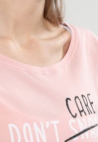 Záběr zblízka osoby ve světle růžovém tričku s částečně viditelným textem „DON'T CARE“ černými a bílými písmeny.